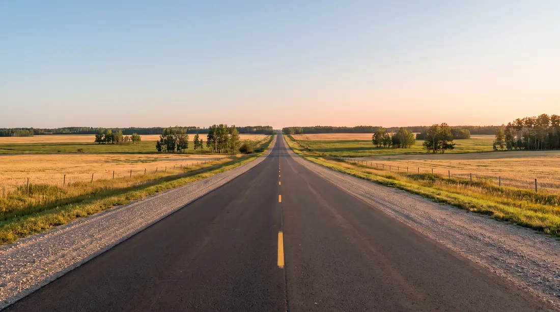 Generated illustration: a straight two-lane highway through flat farmland and aspen bluffs under a wide early-morning sky; not a specific road.