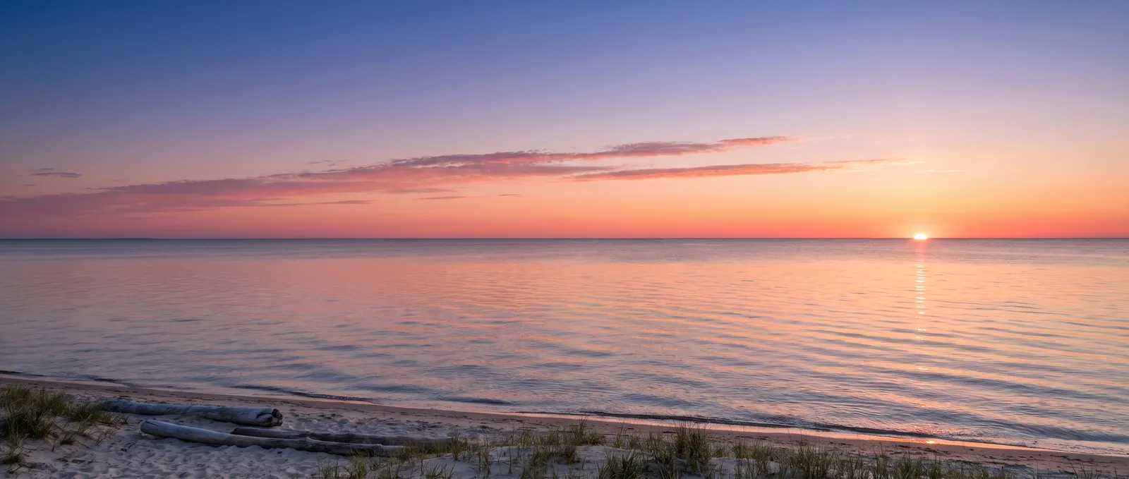 Generated illustration: dawn over a calm lake with a flat horizon across the middle, a small sun on the horizon and a sandy shore with driftwood; not a photograph of the resort.